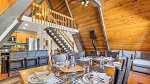 A dining area with a high ceiling, wooden beams, and a staircase leading to an upper level. - Nakai Trail Vacation Rental | 4-BR A-Frame in Kachina Village, Flagstaff | Loft • Firepit  • Game Rm • Sauna • New Bathroom (Flagstaff)