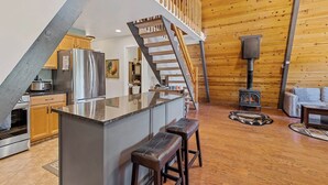 A spacious kitchen with a central island, a wooden staircase, and a cozy seating area with a fireplace. - Nakai Trail Vacation Rental | 4-BR A-Frame in Kachina Village, Flagstaff | Loft • Firepit  • Game Rm • Sauna • New Bathroom (Flagstaff)