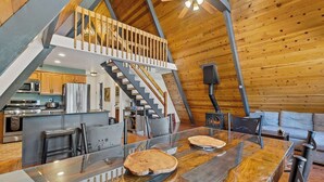 A spacious loft with a high ceiling, a wooden staircase, a kitchen with modern appliances, and a large dining area with a marble countertop. - Nakai Trail Vacation Rental | 4-BR A-Frame in Kachina Village, Flagstaff | Loft • Firepit  • Game Rm • Sauna • New Bathroom (Flagstaff)