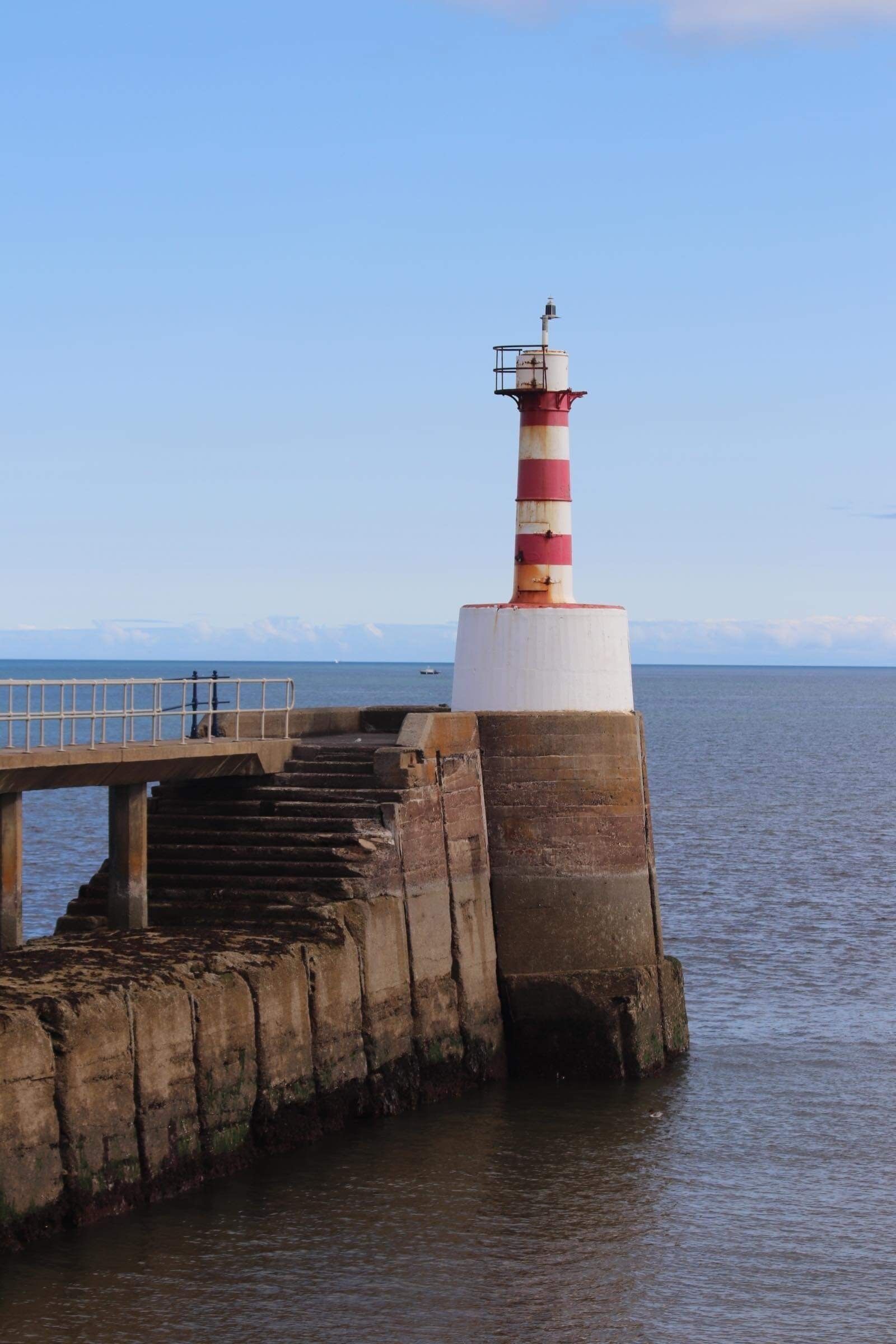 Un faro con la parte superiore a righe rosse e bianche, posto su un molo in cemento che si protende in acque calme.