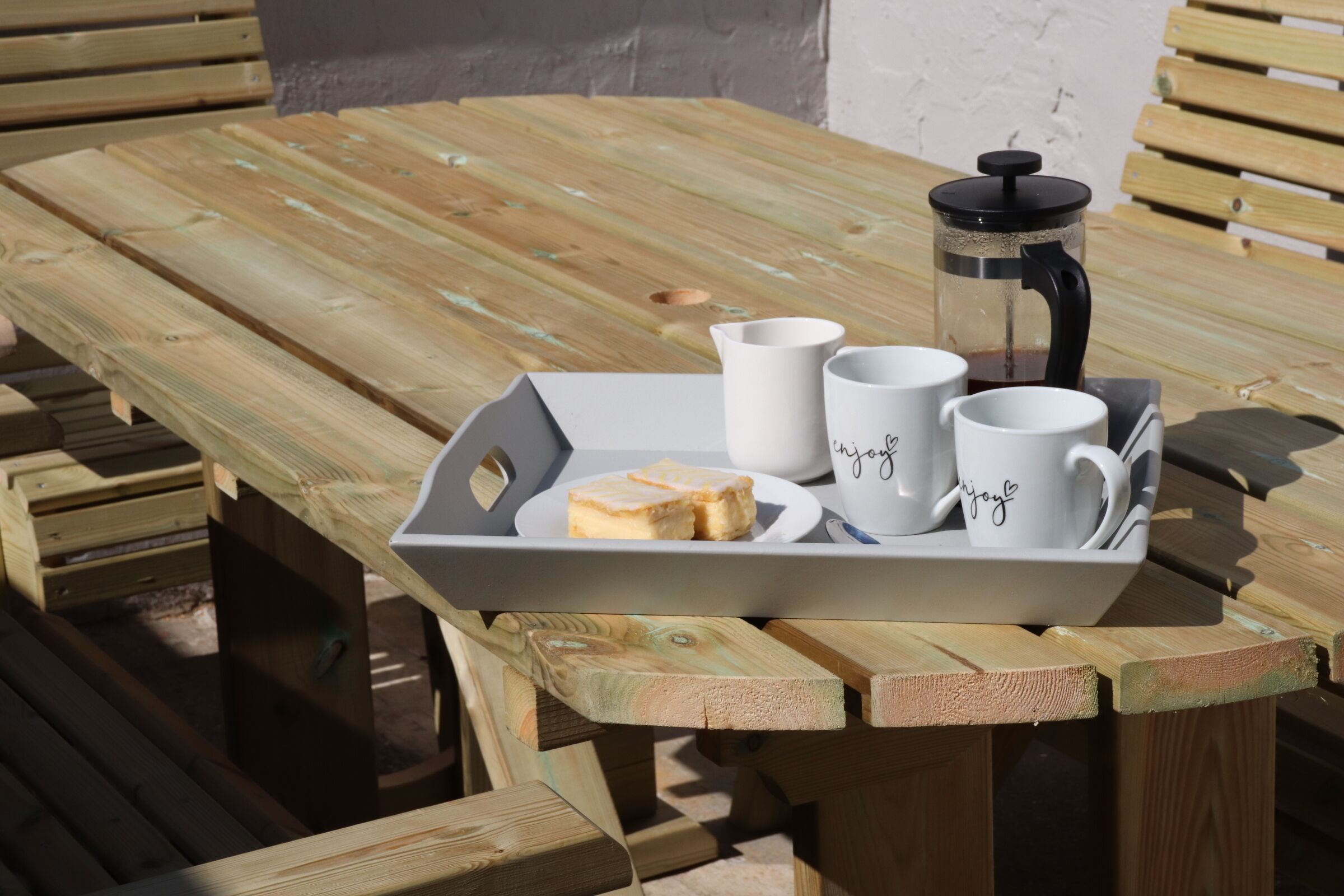 A wooden picnic table with a tray holding two white mugs with the word "joy" and a French press.