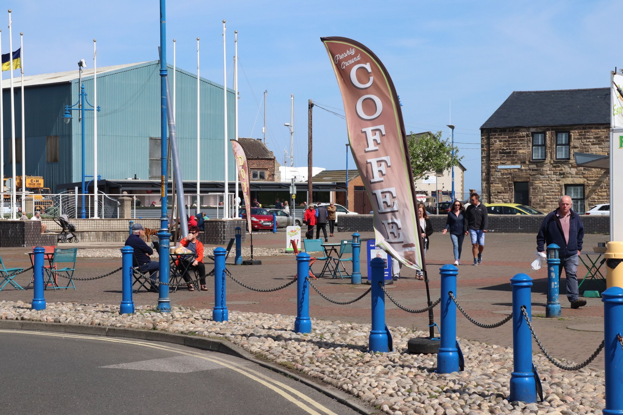 A public square with a large 'COFFEE' banner, stone buildings, and a blue metal fence.