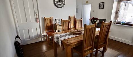 A dining room with a wooden table and chairs, a wall clock, and a window with blinds.