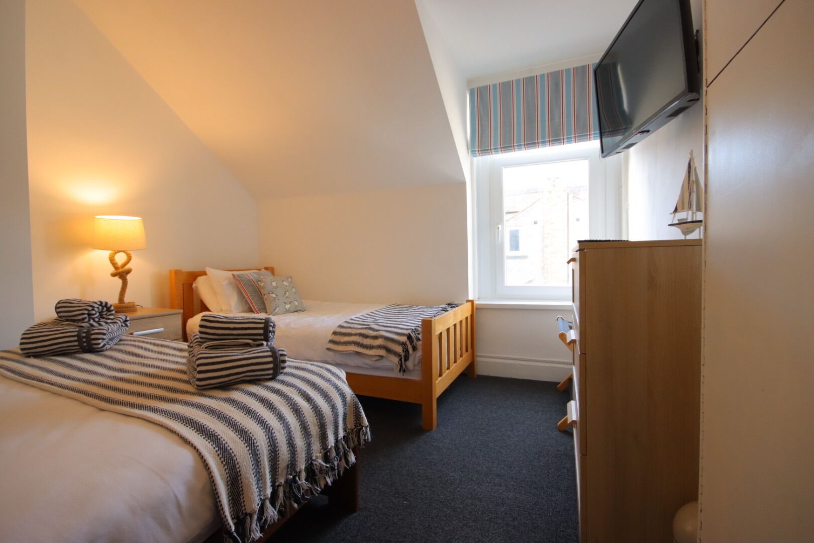 A bedroom with two single beds, a wooden wardrobe, a television mounted on the wall, and a window with a view of buildings outside.