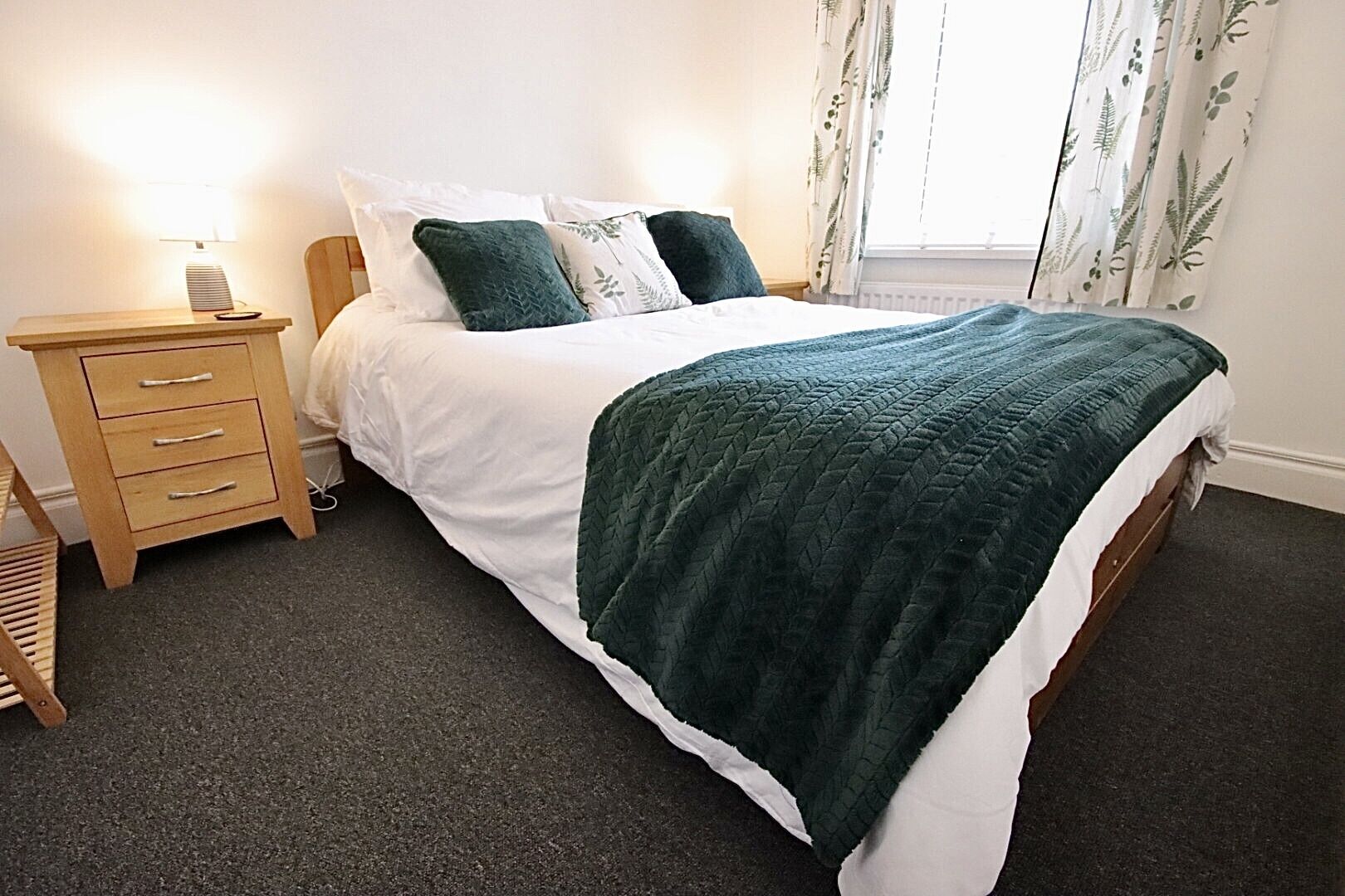 A neatly made bed with a green quilt, a wooden bedside table with drawers, and a lamp.