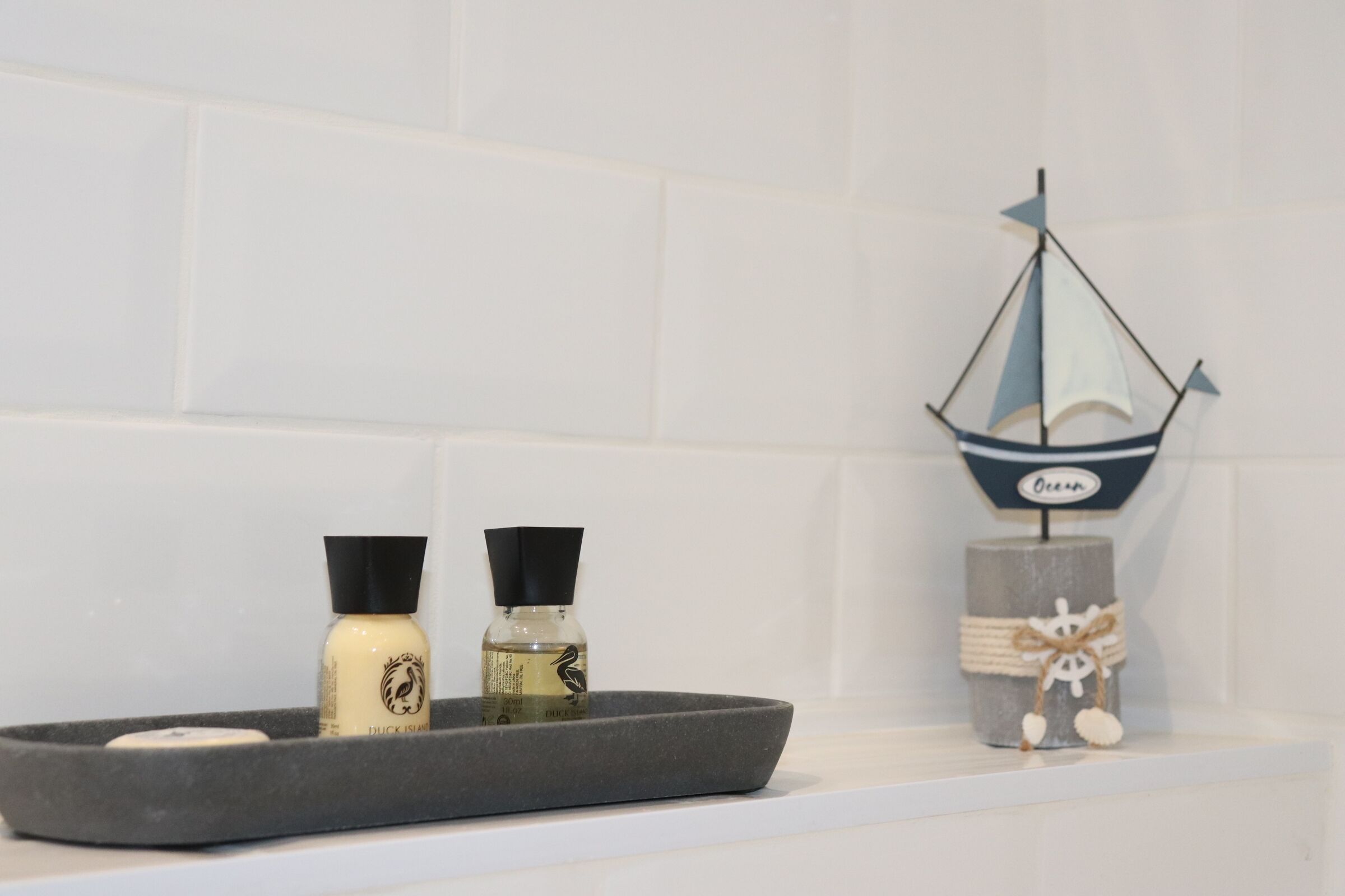 A bathroom with a grey tray holding two bottles, a ship decoration, and a candle on a white shelf.