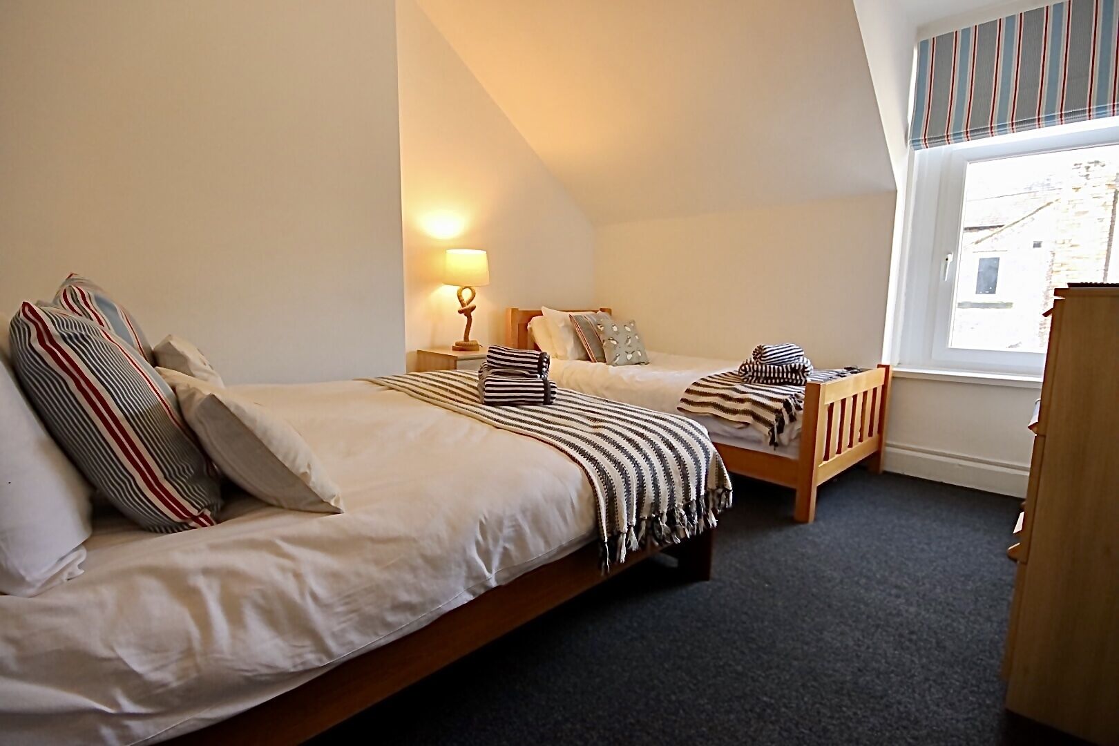 A bedroom with two single beds, a nightstand, a lamp, and a window with a view of a building.
