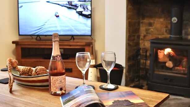 Una sala de estar acogedora con una mesa de madera, una botella de champán, dos copas de vino, un cuenco de galletas, un televisor mostrando una carrera y una chimenea.