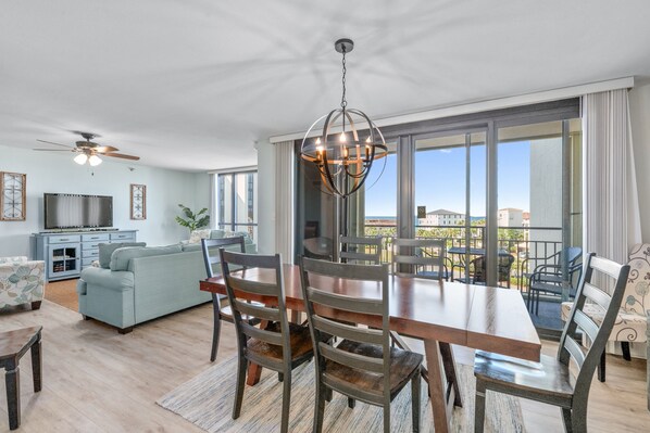 A modern living room with a dining area, large windows, and a balcony. - Steps to the Beach, Pool, Location, Location! Great for Families and Groups (Destin)