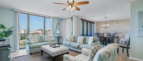 A spacious living room with a ceiling fan, large windows, and a dining area with a chandelier.