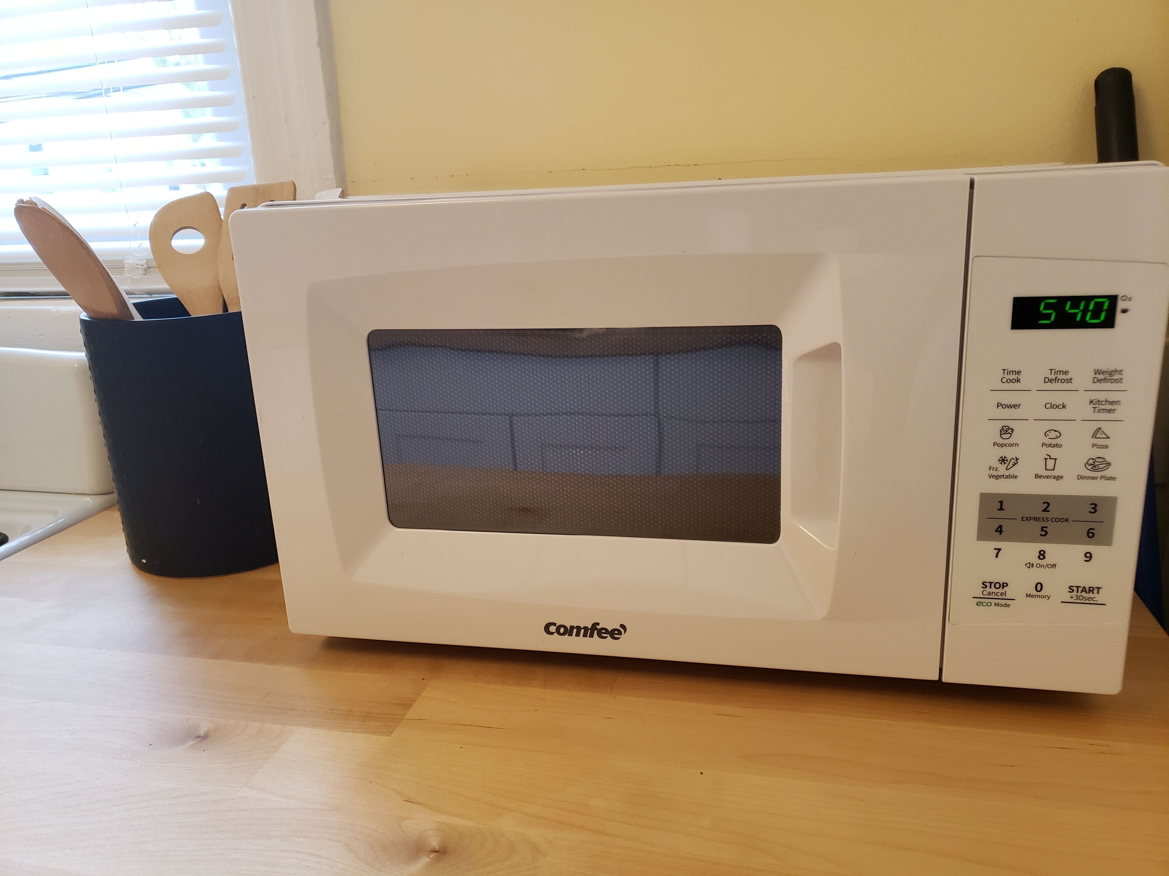 Eine weiße Mikrowelle mit digitalem Display und Bedienfeld, daneben ein Besteckhalter aus Holz.
