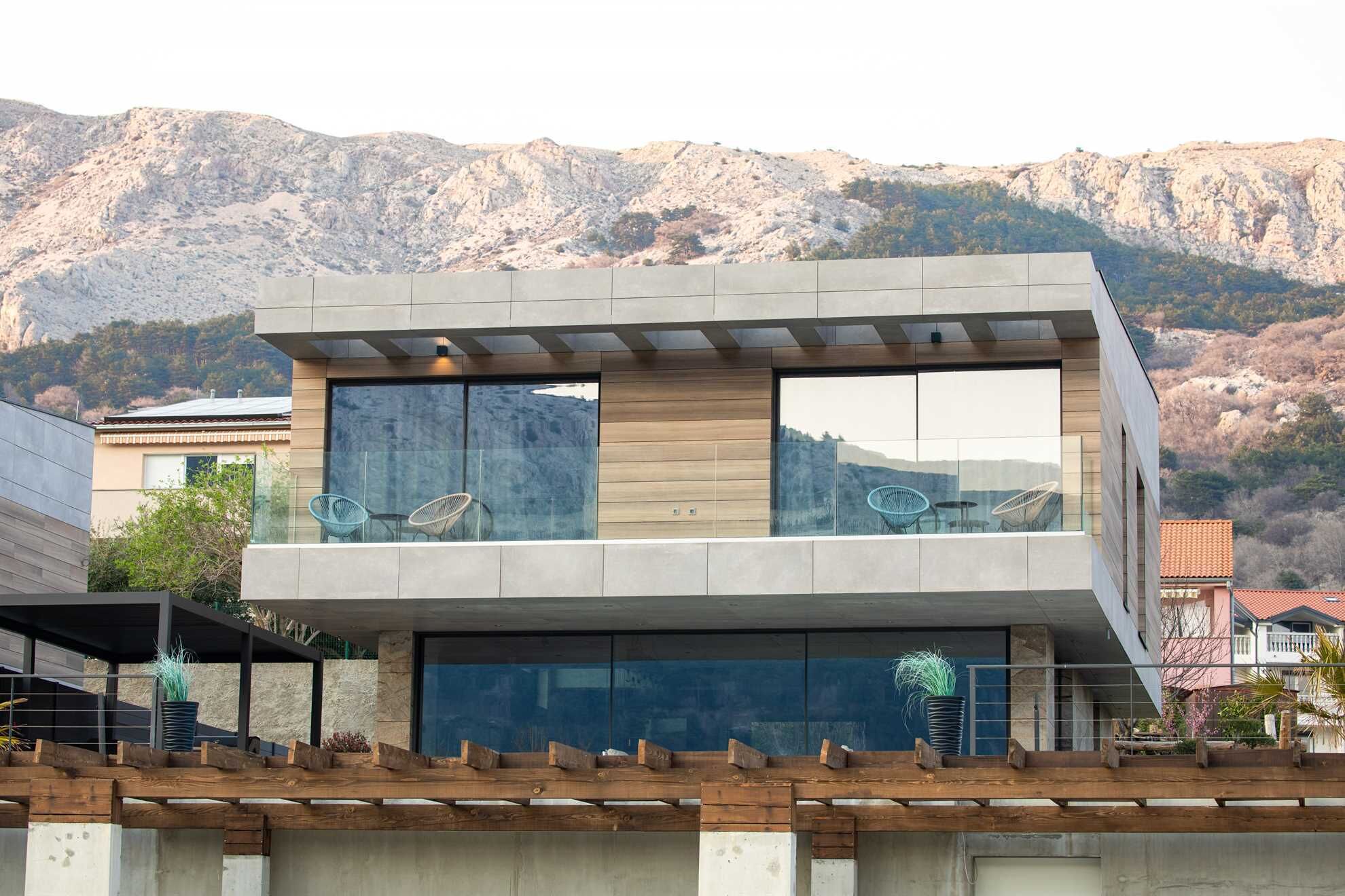 A modern two-story building with large windows, a balcony with chairs, and a view of a mountainous landscape.