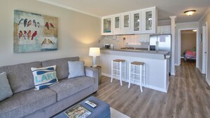 A modern kitchen with white cabinets and a central island, a living area with a grey sofa and a painting of birds, and a hallway leading to a bedroom. - Sweet Lake Get Away (#2) in the Heart of Hot Springs (Hot Springs)