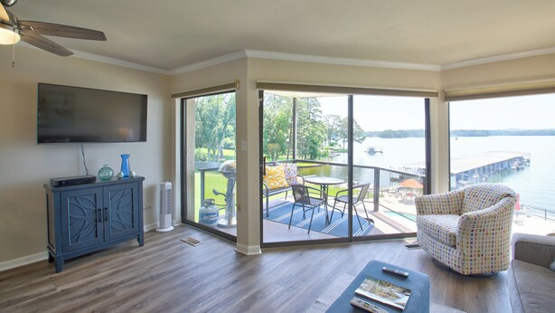 A spacious living room with a large window overlooking a body of water, a ceiling fan, a flat-screen TV, and a seating area with a patterned armchair and a coffee table. - Sweet Lake Get Away (#2) in the Heart of Hot Springs (Hot Springs)