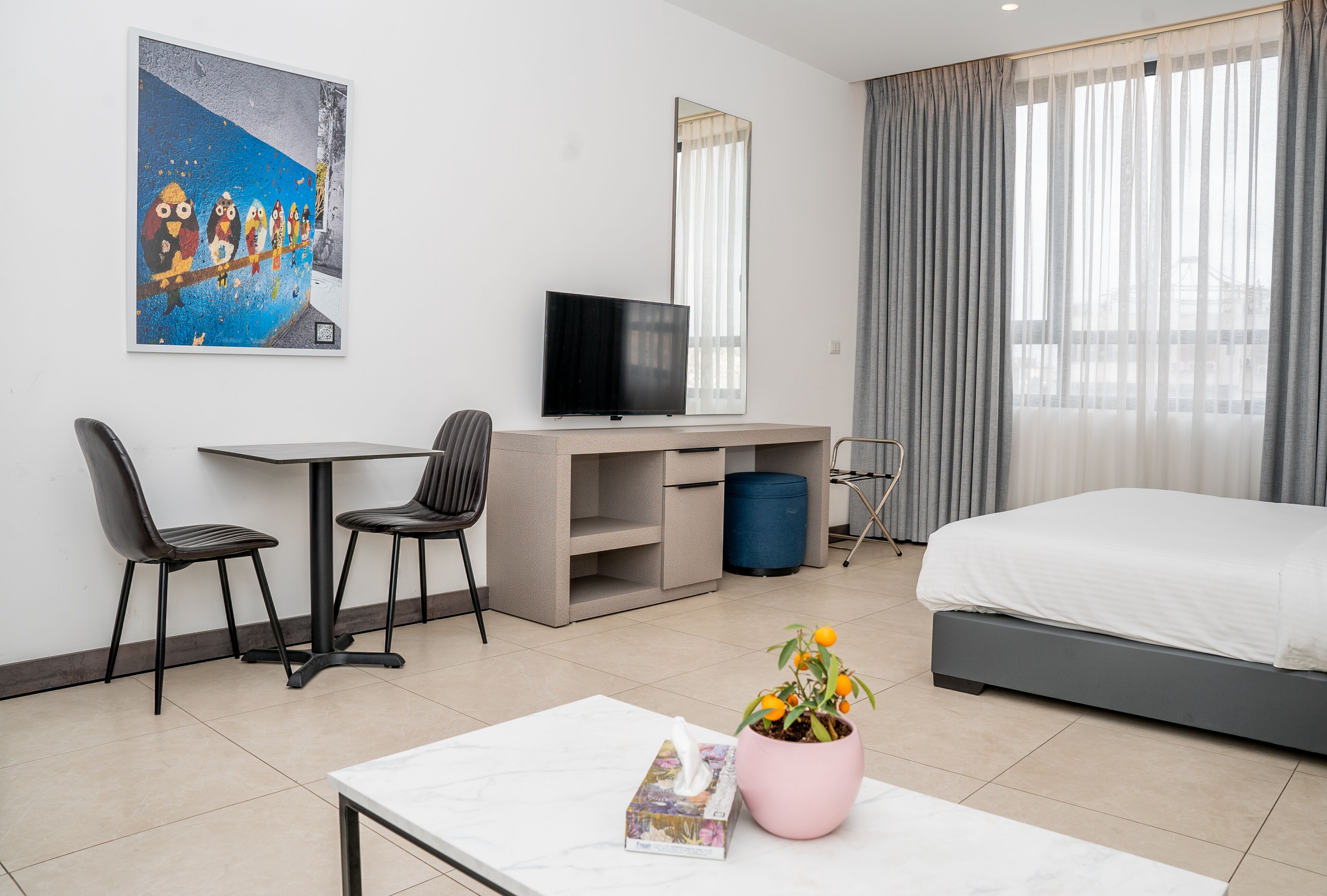 Habitación de hotel con una cama, un escritorio con televisor, dos sillas, una mesa de centro con una revista y una planta, y un ventanal con cortinas.