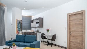 A modern living room with a blue sofa, a yellow ottoman, a small table with a vase of flowers, and a kitchen area with a refrigerator and a sink. - Liv weibdeh (Amman)