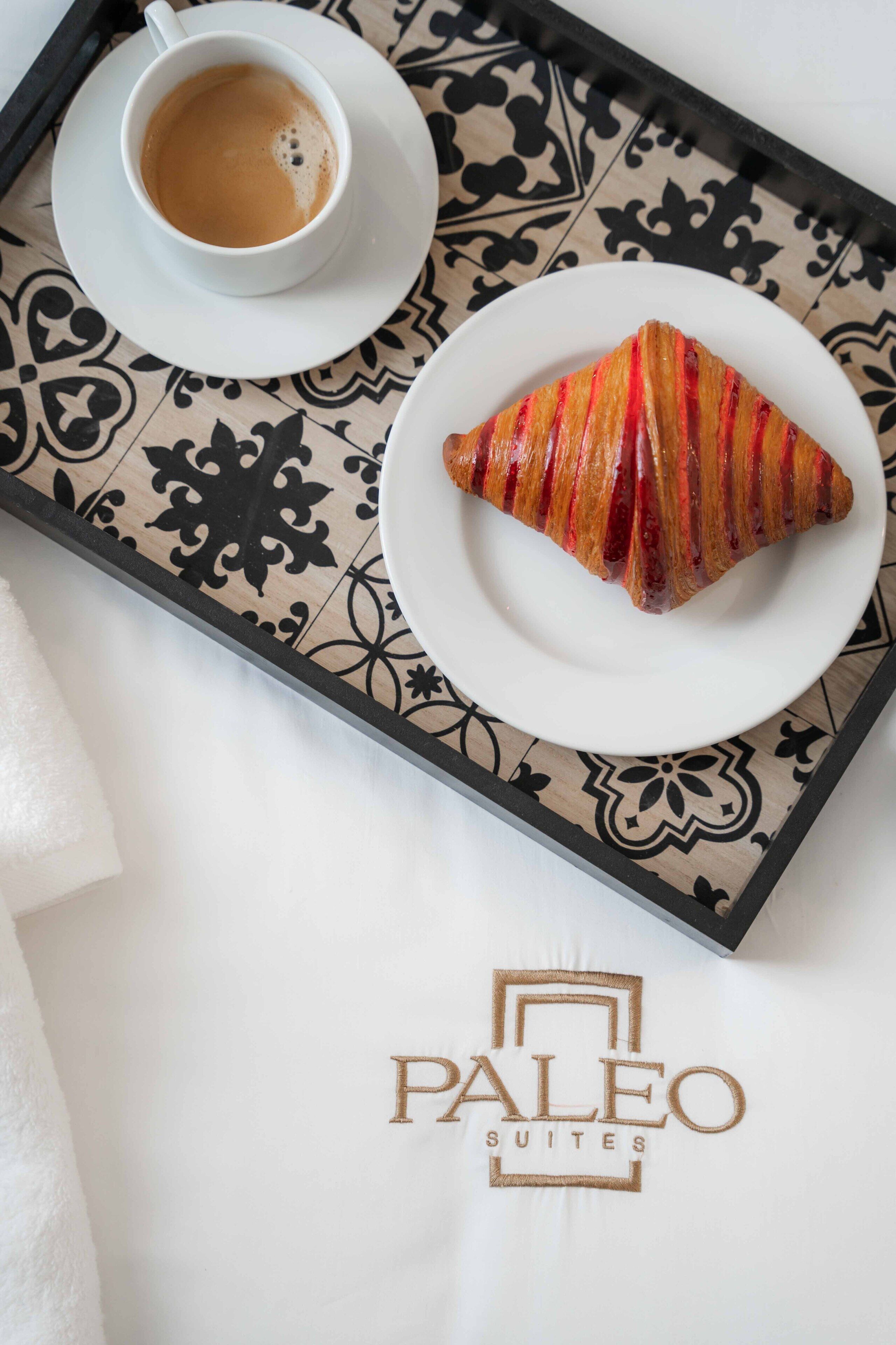 A tray with a cup of coffee and a croissant on a patterned tablecloth.