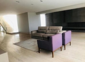 A spacious living room with a grey sofa and two purple armchairs, a large window, and a staircase in the background. - House for rent in the Pampulha region. (Belo Horizonte)