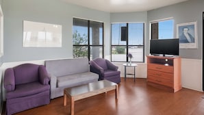 A living room with a grey sofa, purple armchairs, a small coffee table, a TV on a wooden cabinet, and large windows with blinds.