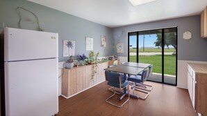 A modern kitchen with a refrigerator, wooden cabinets, a dining table with chairs, and a view through large glass doors.