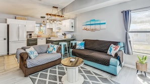 A modern living room with a sofa, coffee table, and kitchen area. - Quaint Cottage by the Bay - Boat Launch 1 Street Over (Elberta)
