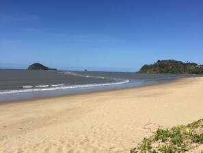 A sandy beach with gentle waves, a small island, and a forested hill in the background.