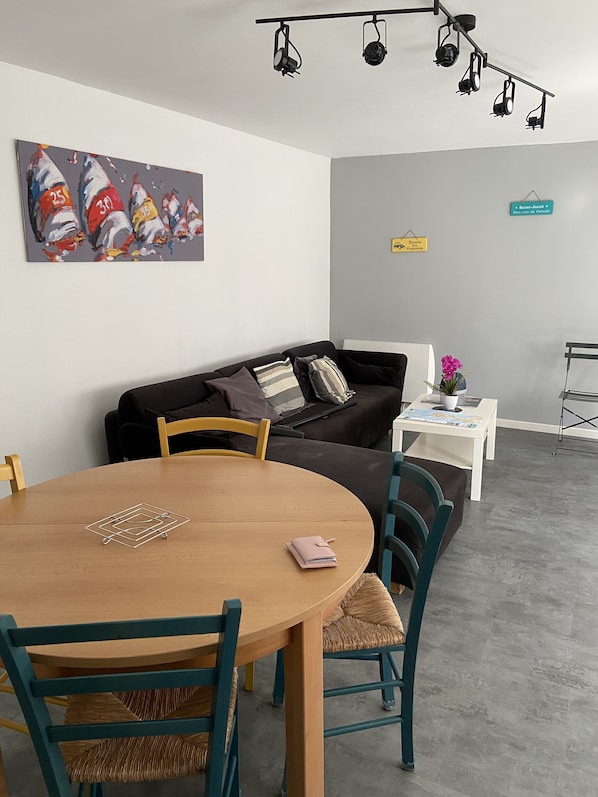 Uma sala moderna com um sofá escuro, uma mesa de jantar de madeira com cadeiras, uma pequena mesa lateral branca com uma planta em vaso e um quadro na parede.