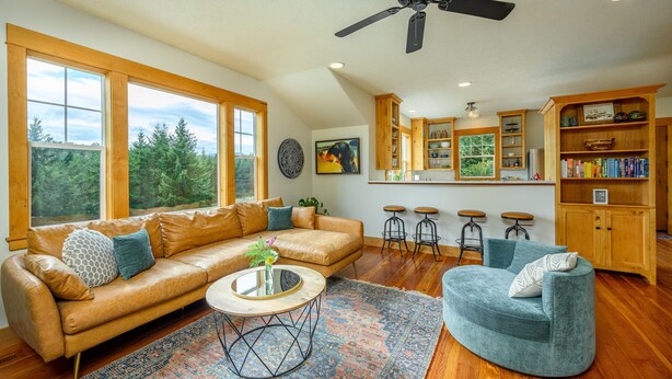 A spacious living room with a leather sofa, a round coffee table, and a teal armchair. There are large windows offering a view of the outdoors, and a kitchen area with wooden cabinets and bar stools. - Rancho Relax Inn  (CARLTON)