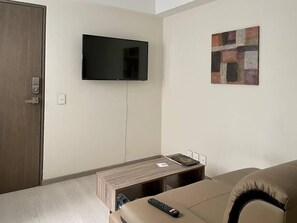 Un salon moderne avec un téléviseur à écran plat fixé au mur, une petite table d’appoint en bois et un canapé beige.