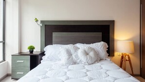 Una cama bien tendida con edredón blanco y varios almohadones, una mesita de noche con lámpara y una pequeña planta en el buró.