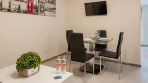 Habitación de hotel con comedor que incluye una mesa de vidrio, sillas negras y un televisor de pantalla plana montado en la pared.
