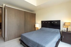 A hotel room with a large wardrobe, a bed with a grey quilt, a bedside table with a lamp, and a small cabinet with bottles.