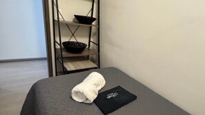 Un lit simple recouvert d’une couverture gris foncé, une serviette blanche roulée et un oreiller noir avec du texte blanc. On aperçoit, à l’arrière-plan, une étagère métallique sur laquelle se trouve un bol, placé sur l’étagère supérieure.