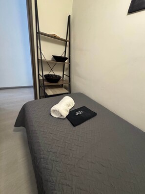 Ein Einzelbett mit dunkelgrauer Bettdecke, ein aufgerolltes weißes Handtuch und ein schwarzes Kopfkissen mit weißer Schrift. Im Hintergrund ist ein Metallregal mit einer Schüssel auf der obersten Ablage zu sehen.