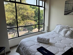 A bedroom with a large window, a bed with white bedding, and a view of trees and parked cars outside.