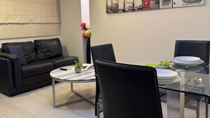 Un salon moderne avec une table à manger en verre, un canapé en cuir noir et une œuvre d’art représentant un paysage urbain accrochée au mur.
