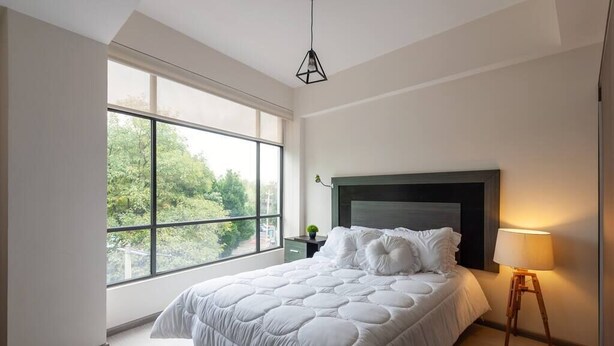A bedroom with a large window, a bed with a white duvet, a bedside table with a lamp, and a view of greenery outside. - Comfortable and reliable stay, convenient location (Mexico City)