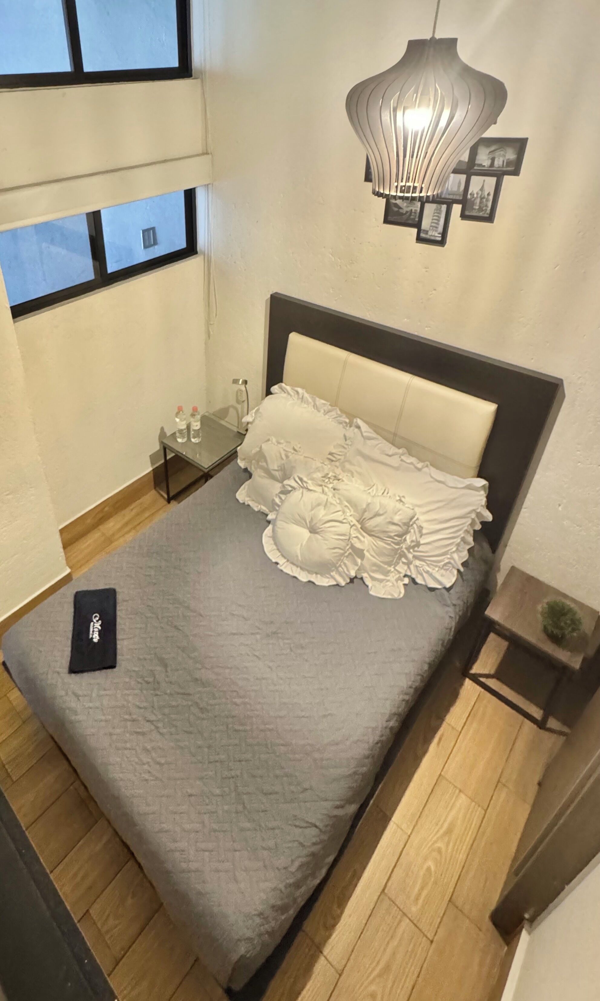 Una cama bien hecha con colcha gris y almohadas con volantes, una mesita de noche con una botella de agua y una lámpara colgante original sobre ella.