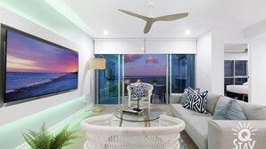 A modern living room with a large TV, comfortable seating, a ceiling fan, and a view of the city through large windows. - 3 Bed Executive Sub Penthouse with Ocean Views (Surfers Paradise)