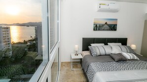 Une chambre d’hôtel avec une grande fenêtre donnant sur un plan d’eau et offrant une vue sur le coucher de soleil. On y trouve un lit avec une literie à motifs et une tête de lit, une petite table de chevet avec une lampe, et un appareil de climatisation fixé au mur.
