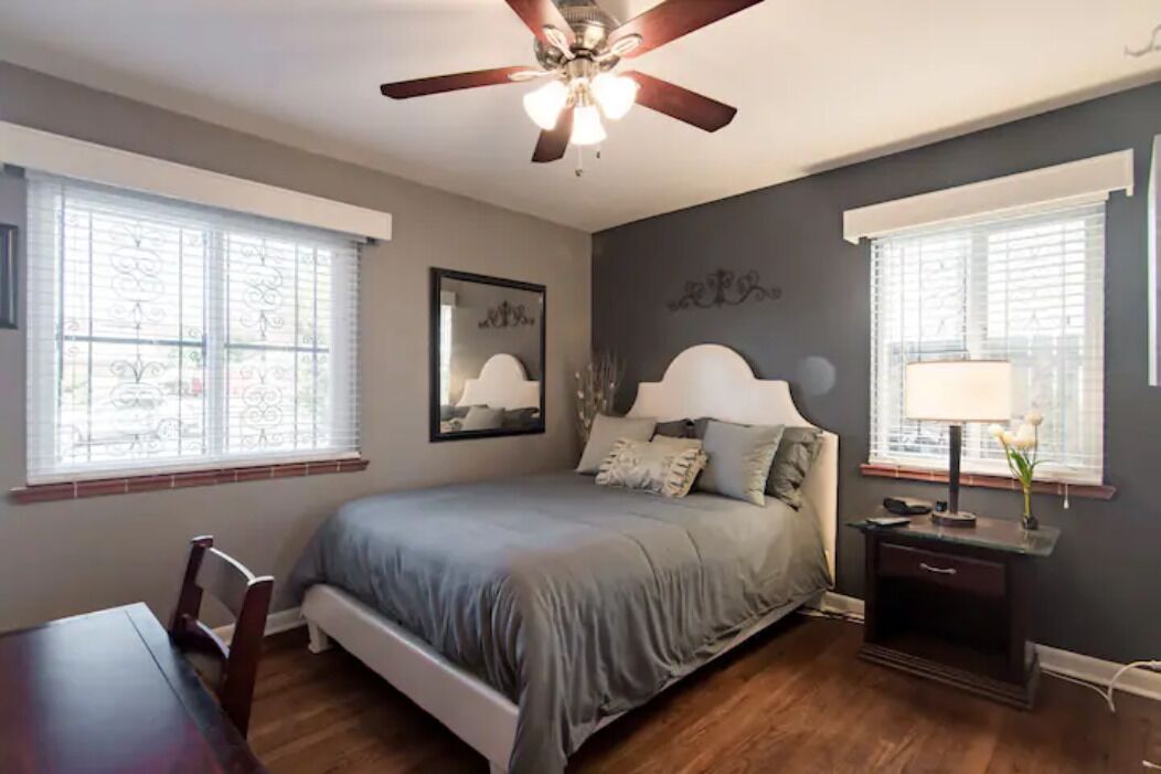 A bedroom with a bed, a desk, a chair, a ceiling fan, and two windows with blinds.