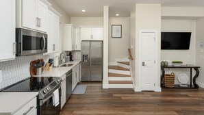 A modern kitchen with white cabinets, stainless steel appliances, and a wooden staircase leading to a living area with a flat-screen TV. - New - Lookout Views 2 Br/2BA Sleeps 8 next to OddStory Brewery! Pet friendly! (Chattanooga)