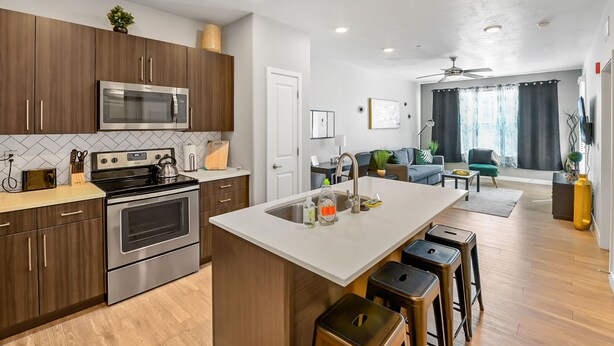 A modern kitchen with stainless steel appliances, wooden cabinets, and a central island with stools. - Experience South Valley charm, just a 9-min drive to Thanksgiving Point (Draper)