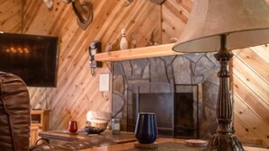 Une pièce chaleureuse avec une cheminée en pierre, un plafond en bois et une table sur laquelle se trouvent une lampe et une tasse bleue.