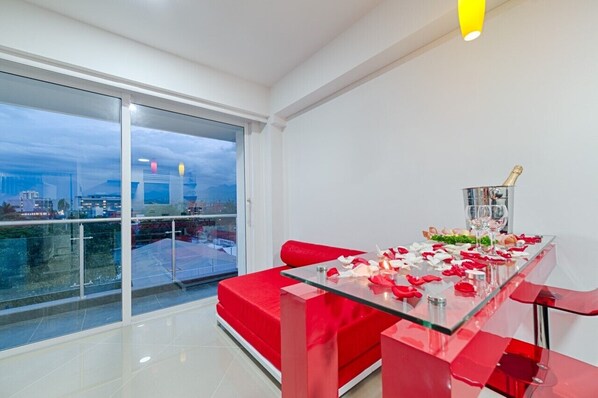 Una sala de estar moderna con una mesa de vidrio para dos personas, muebles rojos y vistas a la ciudad a través de ventanas amplias.