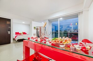 Habitación de hotel moderna con una mesa de vidrio preparada para una cena romántica, una cama con ropa de cama roja y vistas a la ciudad a través de grandes ventanales.