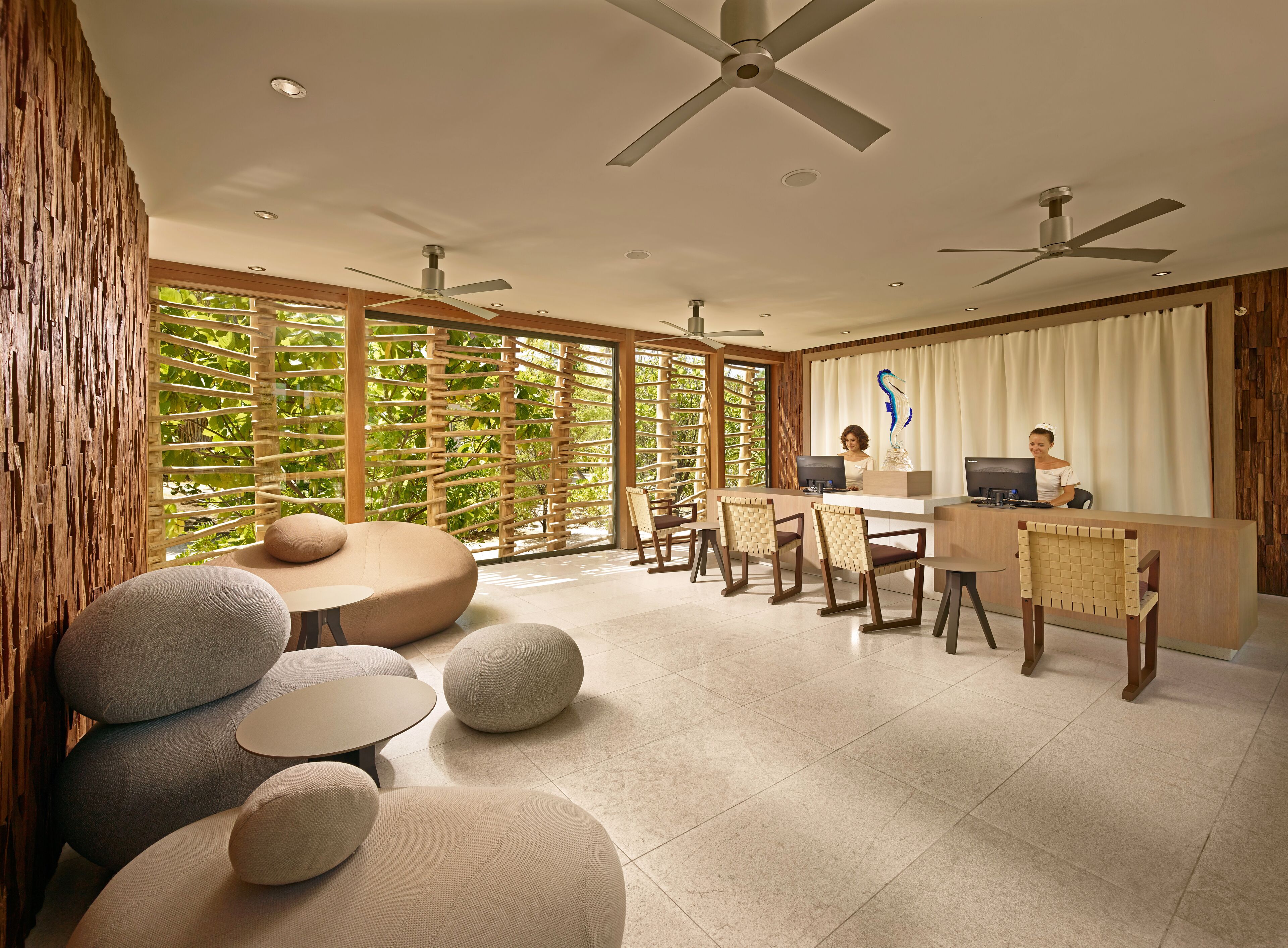 A modern hotel lobby with a reception desk, seating area with round tables and chairs, large windows with a view of greenery, and ceiling fans.