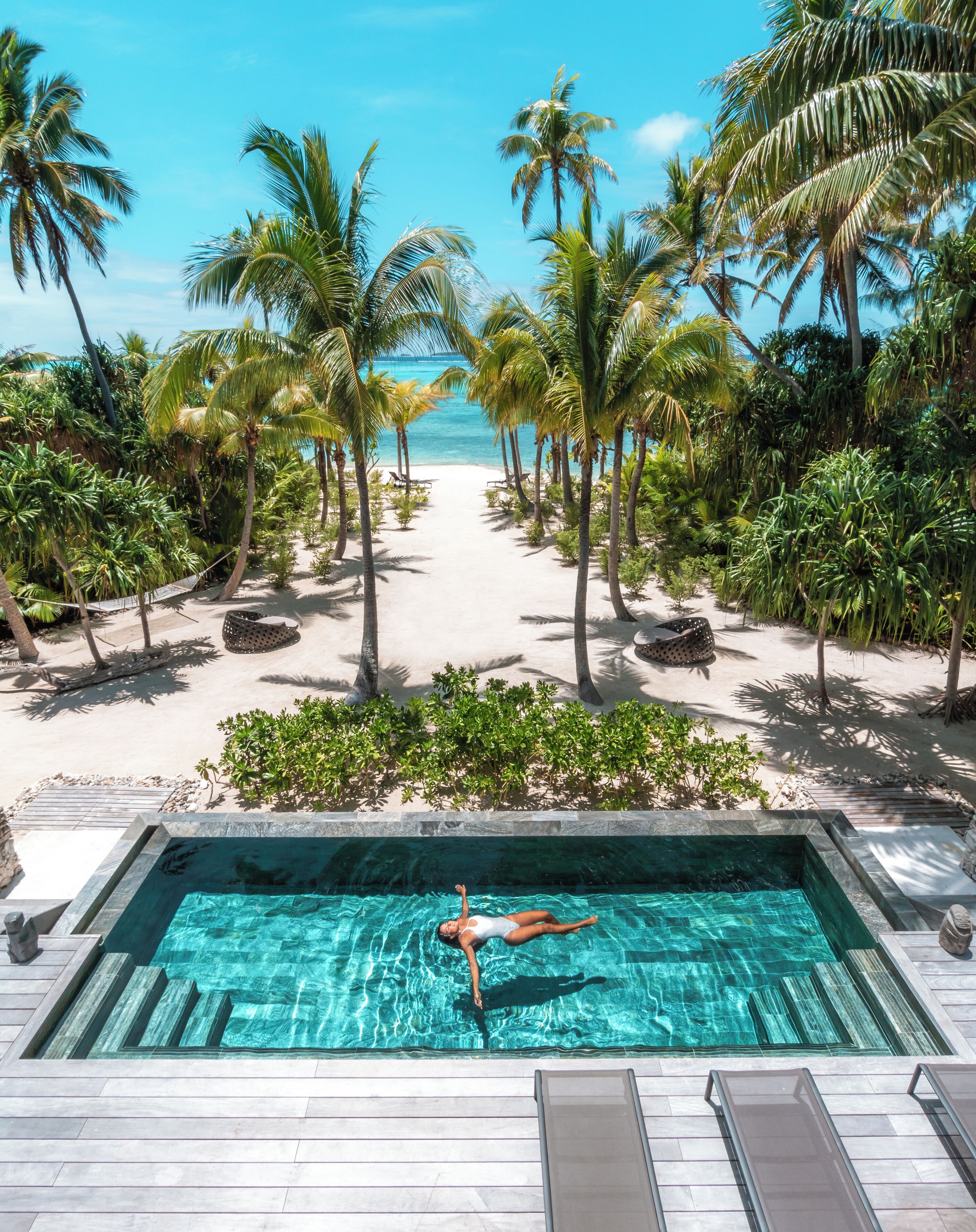 Piscina con una persona flotando, rodeada de palmeras y vista a la playa.