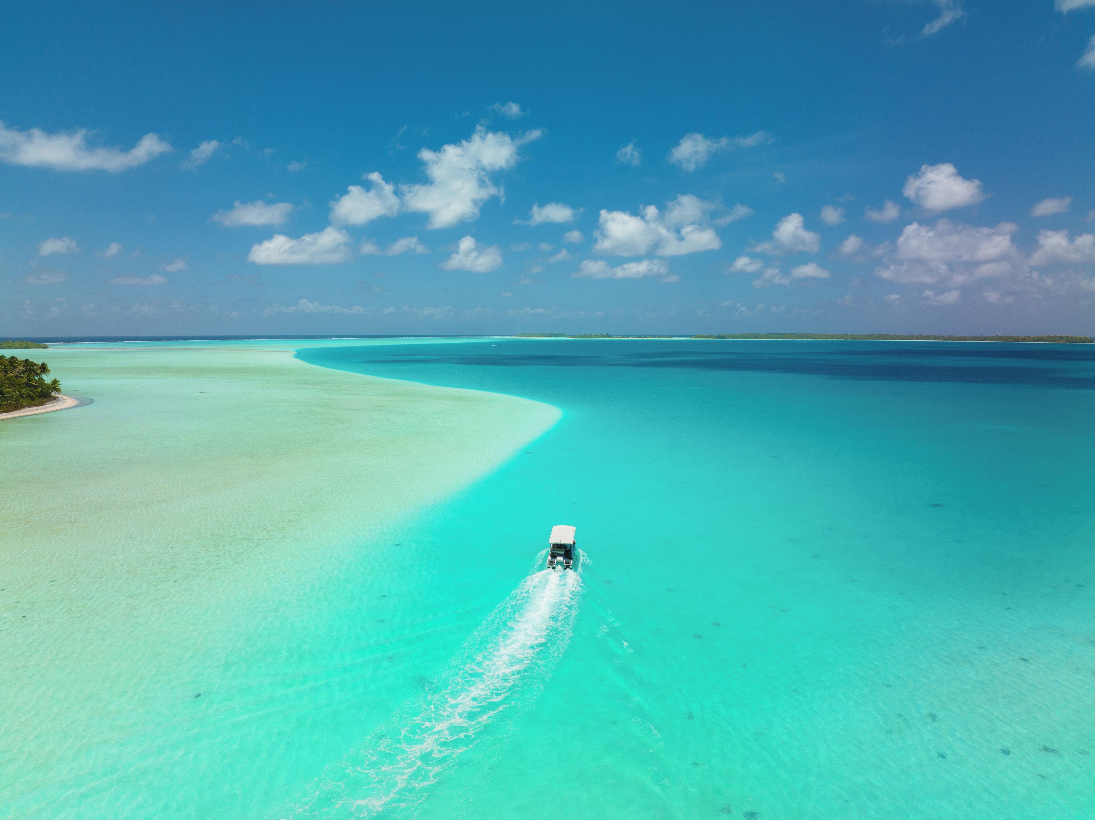 An aerial view of a turquoise sea with a boat creating a wake, surrounded by clear skies and distant land.