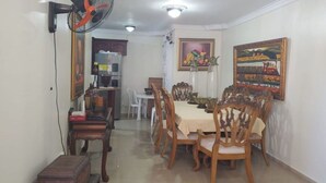 Un coin repas avec une table dressée pour quatre personnes, entourée de chaises en bois. Un ventilateur est fixé au mur, et deux tableaux ornent les murs.